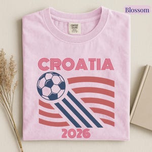 May include: A light pink t-shirt featuring the text CROATIA in red, a graphic of a soccer ball, three diagonal navy blue stripes, red horizontal wavy lines, and the number 2026 in red. The shirt has a Comfort Colors brand label at the neck.