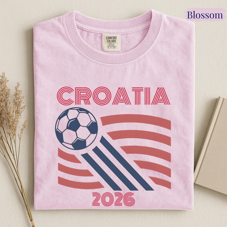 May include: A light pink t-shirt featuring the text CROATIA in red, a graphic of a soccer ball, three diagonal navy blue stripes, red horizontal wavy lines, and the number 2026 in red. The shirt has a Comfort Colors brand label at the neck.