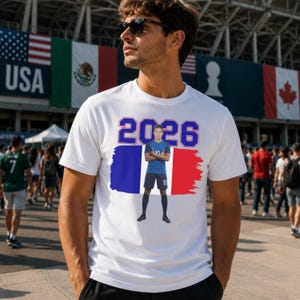 Może przedstawiać: Biała koszulka z grafiką przedstawiającą niebieską liczbę "2026", niebiesko-biało-czerwony wz&oacute;r i piłkarza. Koszulka jest noszona przez osobę noszącą okulary przeciwsłoneczne.