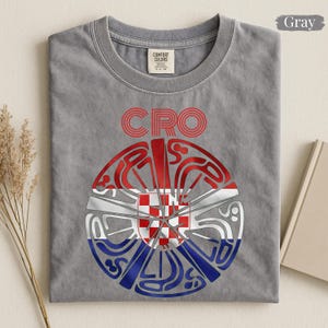 May include: A gray crew neck t-shirt featuring the text CRO in red above a circular graphic. The graphic contains a red and white checkered pattern in the center, surrounded by stylized red, white, and blue lines forming a circular shape.