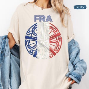 May include: An ivory t-shirt featuring the text FRA above a circular graphic. The circle contains a stylized design with blue, white, and red segments resembling the French flag, overlaid with intricate, abstract silver-toned line patterns.