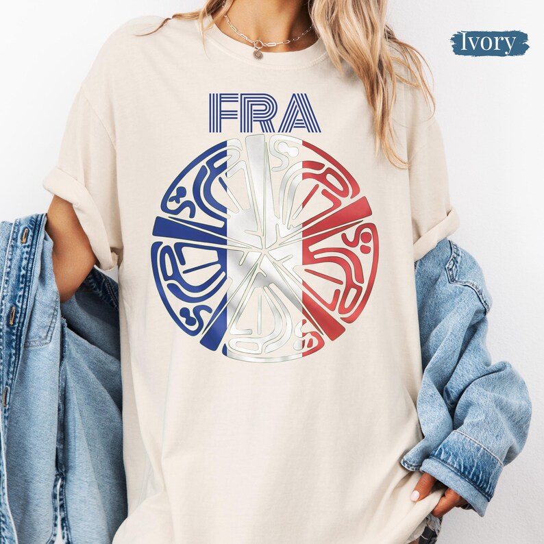 May include: An ivory t-shirt featuring the text FRA above a circular graphic. The circle contains a stylized design with blue, white, and red segments resembling the French flag, overlaid with intricate, abstract silver-toned line patterns.