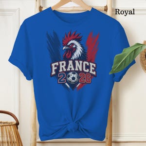 May include: Royal blue t-shirt featuring a graphic of a rooster with red, white, and blue accents, the word "FRANCE", the numbers "2026", and a soccer ball. The shirt is displayed on a wooden hanger.