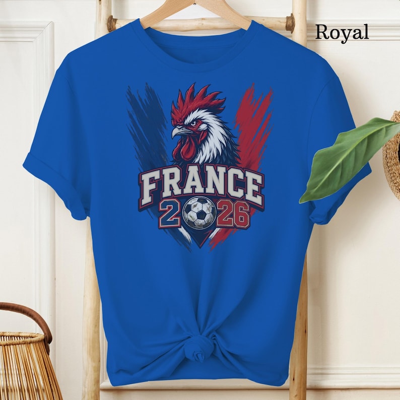 May include: Royal blue t-shirt featuring a graphic of a rooster with red, white, and blue accents, the word "FRANCE", the numbers "2026", and a soccer ball. The shirt is displayed on a wooden hanger.