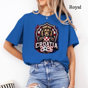 May include: Royal blue t-shirt featuring a graphic of a lion holding a Croatian checkered shield, with the text "CROATIA 2026" and a soccer ball. The design is framed by red and blue accents.