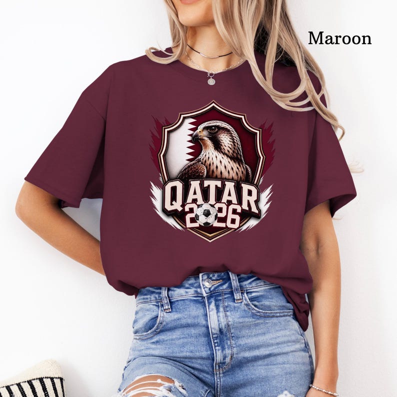 May include: Maroon t-shirt featuring a graphic of a falcon inside a shield with the Qatar flag and the text "QATAR 2026". The shirt is a solid maroon color and has short sleeves. The design includes a soccer ball.