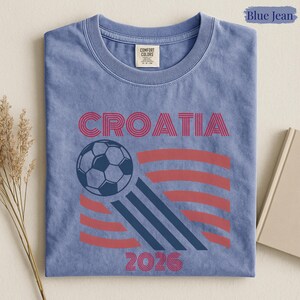May include: A blue jean colored t-shirt featuring the text CROATIA in red, a graphic of a soccer ball, three diagonal dark blue stripes, and the year 2026 in red. The shirt has a crew neckline and a Comfort Colors brand label at the inner neck.