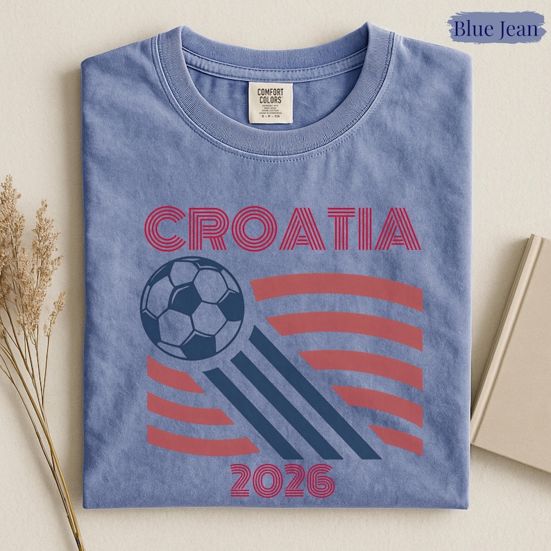 May include: A blue jean colored t-shirt featuring the text CROATIA in red, a graphic of a soccer ball, three diagonal dark blue stripes, and the year 2026 in red. The shirt has a crew neckline and a Comfort Colors brand label at the inner neck.