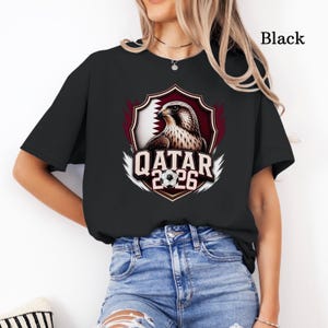 May include: Black t-shirt featuring a graphic of a falcon, the Qatar flag, and the text "QATAR 2026". The design is set within a shield shape with a soccer ball. The text "Black" is printed on the upper right.
