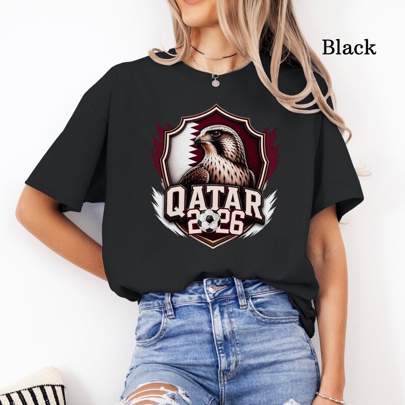 May include: Black t-shirt featuring a graphic of a falcon, the Qatar flag, and the text "QATAR 2026". The design is set within a shield shape with a soccer ball. The text "Black" is printed on the upper right.