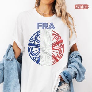 May include: A white t-shirt featuring the text FRA in blue, positioned above a circular graphic. The circle is divided into three vertical segments colored blue, white, and red, with stylized, abstract line art patterns overlaid across the design.