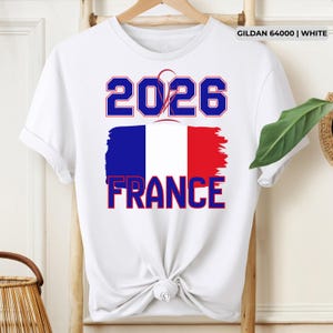 May include: White t-shirt with the text "2026 FRANCE" in blue and red, featuring a stylized French flag design. The shirt is made of white fabric and has a crew neck.