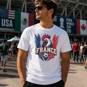 May include: White t-shirt featuring a graphic of a rooster in red, white, and blue, with the text "FRANCE 2026" and a soccer ball. The shirt is short-sleeved.