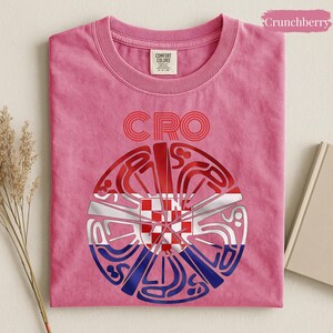 May include: A pink short-sleeved t-shirt featuring the text CRO above a circular graphic. The graphic contains a stylized red and white checkerboard pattern overlaid with blue and red curved lines. The shirt has a Comfort Colors brand label.