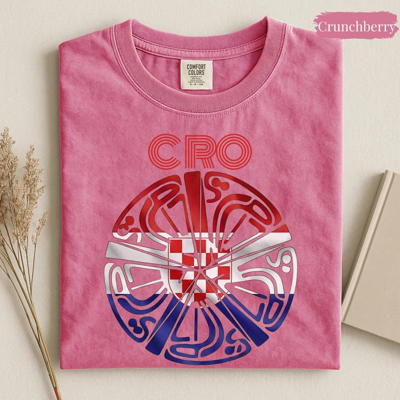 May include: A pink short-sleeved t-shirt featuring the text CRO above a circular graphic. The graphic contains a stylized red and white checkerboard pattern overlaid with blue and red curved lines. The shirt has a Comfort Colors brand label.