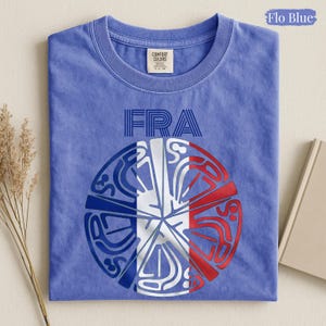May include: A Flo Blue t-shirt featuring the text FRA above a circular graphic. The design incorporates the blue, white, and red colors of the French flag within a stylized, segmented pattern. The shirt has a crew neckline and a visible brand label.