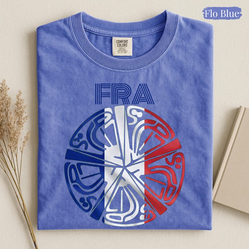 May include: A Flo Blue t-shirt featuring the text FRA above a circular graphic. The design incorporates the blue, white, and red colors of the French flag within a stylized, segmented pattern. The shirt has a crew neckline and a visible brand label.