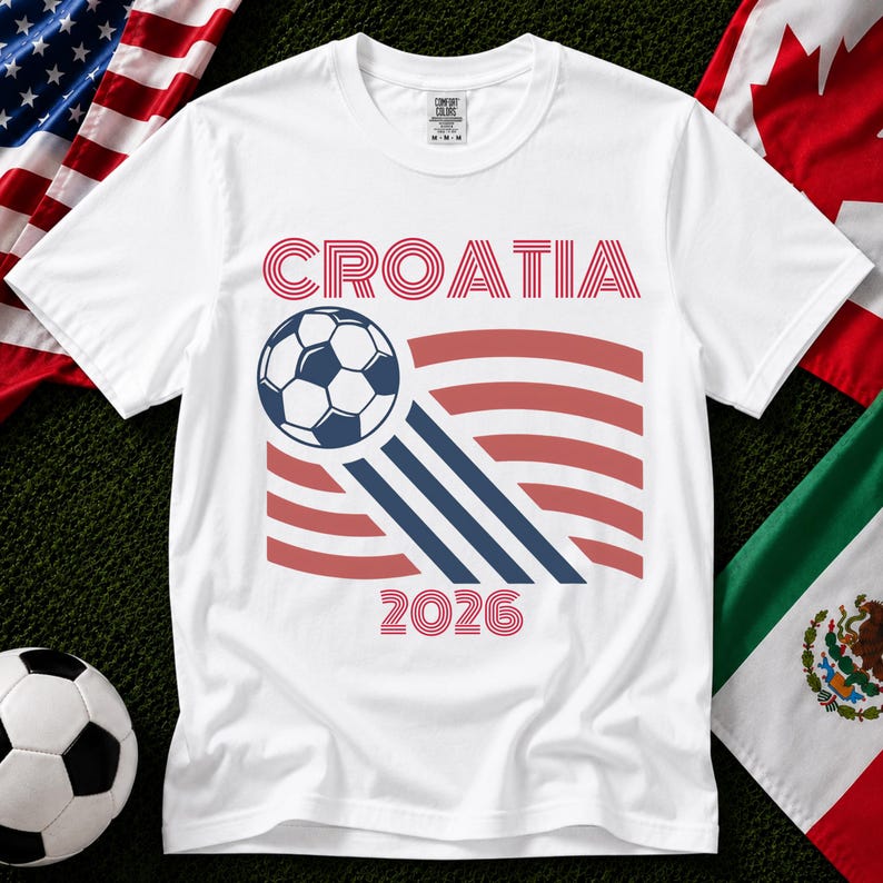 May include: A white short-sleeve t-shirt featuring the text CROATIA in red, a graphic of a soccer ball with blue motion lines, and the year 2026 in red. The design includes red curved stripes behind the soccer ball on the front of the shirt.