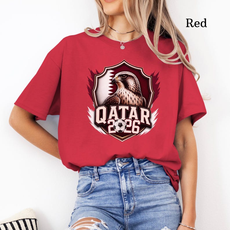 May include: Red t-shirt featuring a shield design with a falcon, the Qatar flag, and the text "QATAR 2026" in white. The shirt is a solid red color.