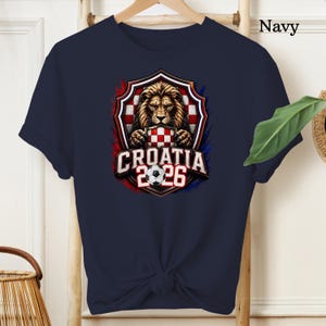 May include: Navy blue t-shirt featuring a detailed graphic of a lion holding a Croatian checkered emblem, with the text "CROATIA 2026" and a soccer ball. The design is framed by red and blue accents.