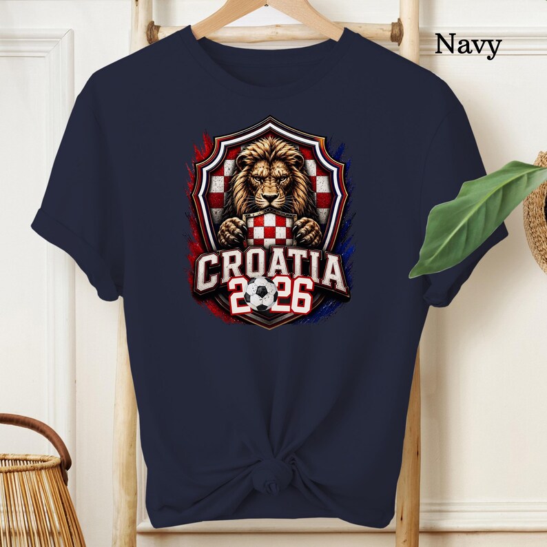 May include: Navy blue t-shirt featuring a detailed graphic of a lion holding a Croatian checkered emblem, with the text "CROATIA 2026" and a soccer ball. The design is framed by red and blue accents.