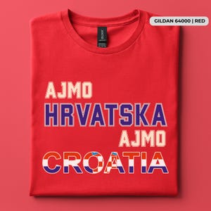 May include: A red short-sleeved t-shirt featuring the text AJMO, HRVATSKA, AJMO, and CROATIA printed in a bold, block-style font. The word CROATIA incorporates a red and white checkered pattern within the letters. A label reads GILDAN 64000 | RED.