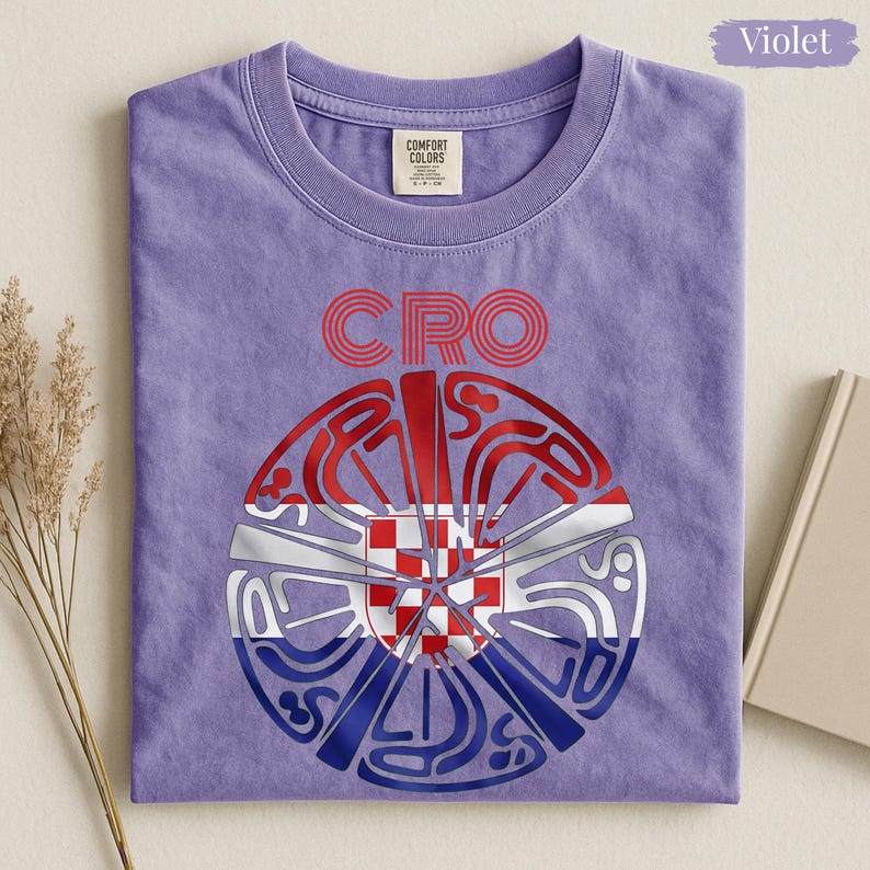 May include: A violet crew neck t-shirt featuring the text CRO above a circular graphic. The graphic contains a red, white, and blue pattern with a central red and white checkered shield design. The shirt has a Comfort Colors brand label at the neck.