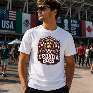 May include: White t-shirt featuring a shield design with a lion holding a checkered pattern, the text "CROATIA 2026", and a soccer ball. The design is in red, white, and blue colors. The shirt is worn by a person wearing sunglasses.