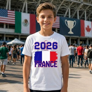 May include: A white t-shirt with the text "2026 FRANCE" in blue and red, featuring the French flag design. The shirt is worn by a person standing in front of a crowd and flags.