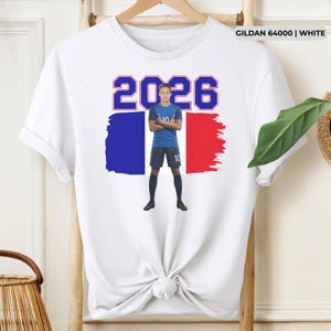 Może przedstawiać: Biała koszulka z grafiką piłkarza w niebieskim stroju, numer 10 i rokiem 2026. Projekt zawiera kolory francuskiej flagi: niebieski, biały i czerwony. Koszulka to Gildan 64000.