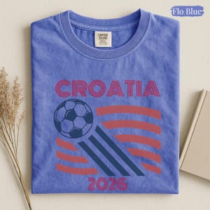 May include: A Flo Blue short-sleeve t-shirt with a crew neckline. The graphic features the word CROATIA in red, a soccer ball, and the year 2026 in red. The design includes red and dark blue curved lines. The shirt has a Comfort Colors label.