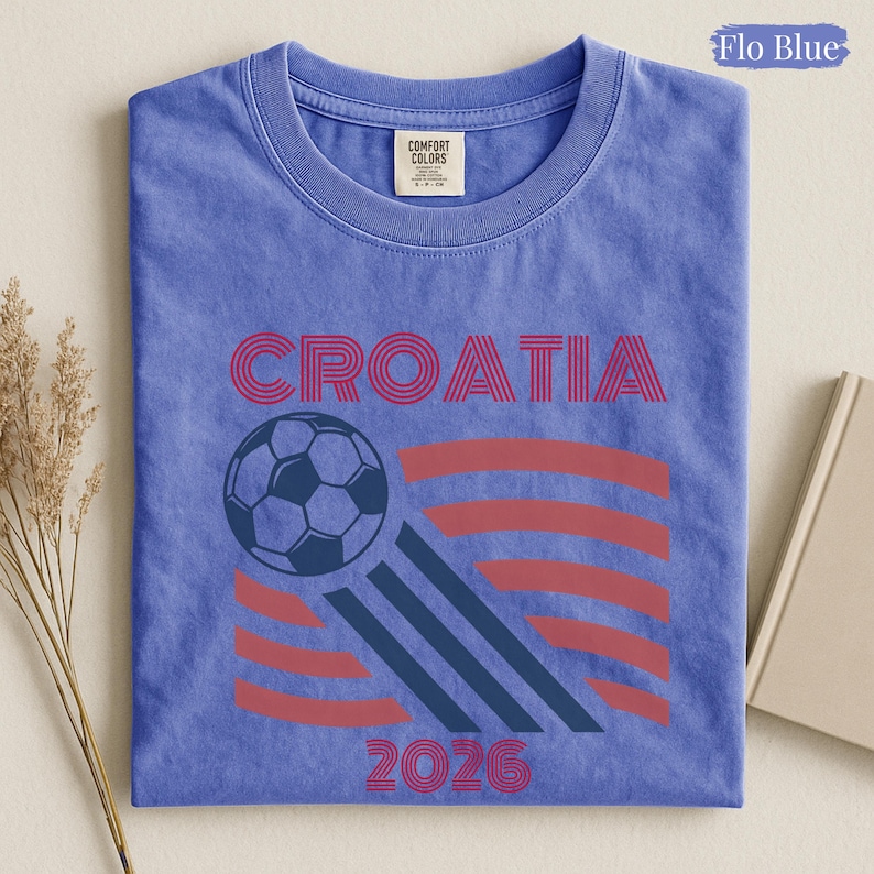 May include: A Flo Blue short-sleeve t-shirt with a crew neckline. The graphic features the word CROATIA in red, a soccer ball, and the year 2026 in red. The design includes red and dark blue curved lines. The shirt has a Comfort Colors label.