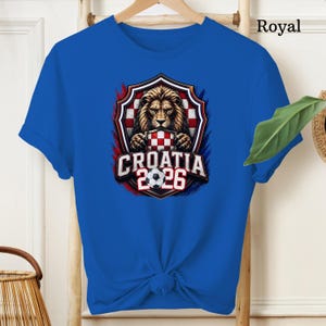May include: Royal blue t-shirt featuring a graphic of a lion holding a Croatian flag, with the text "CROATIA 2026" and a soccer ball. The design is set within a shield shape with red, white, and blue accents.