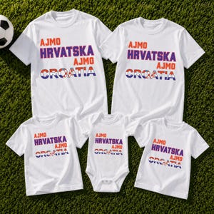 May include: A set of five white t-shirts and one white baby bodysuit, each featuring the orange text AJMO, purple text HRVATSKA, and the word CROATIA in blue, white, and red, with a soccer ball graphic integrated into the letter O in Croatia.