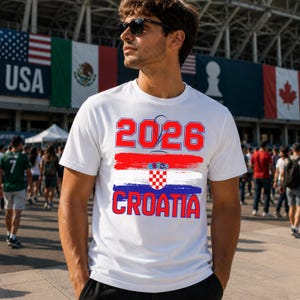 May include: White t-shirt with the text "2026 CROATIA" in red and blue. The design includes a Croatian flag graphic. The shirt is worn by a person wearing sunglasses, with flags in the background.
