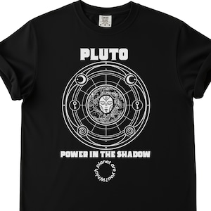 May include: Black t-shirt with the word "PLUTO" in white block letters above a circular graphic. The graphic features a detailed white design with a central face and the text "POWER IN THE SHADOW". The shirt also has the text "Which planet are you?" in a circle.