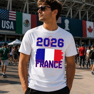May include: White t-shirt with the text "2026 FRANCE" in blue and red, featuring the French flag design. The shirt is worn by a person wearing sunglasses, with flags of USA, Mexico, and Canada in the background.