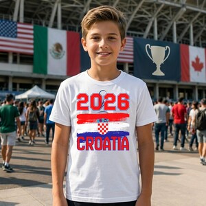 May include: White t-shirt with the text "2026" in red, a red and white stripe, the Croatian flag, and the word "CROATIA" in red. The shirt is worn by a person in front of a crowd and flags.