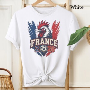 May include: White t-shirt featuring a graphic of a rooster with the French flag colors and the text "FRANCE 2026" and a soccer ball. The shirt is tied at the waist.