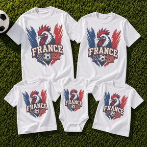 May include: White t-shirts and a onesie with a graphic featuring a rooster, the text "FRANCE 2026", and a soccer ball. The design includes blue and red brushstrokes, resembling the French flag.