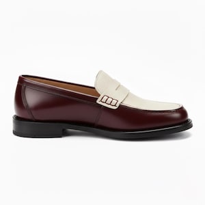 May include: A two-tone penny loafer featuring a smooth, deep burgundy leather heel and side panel contrasted with a textured, off-white leather vamp and saddle strap. The shoe is finished with a solid black sole and a low, stacked heel.