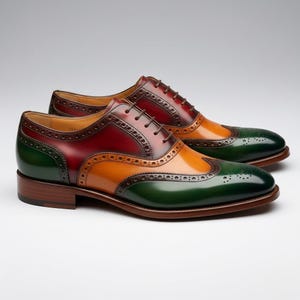 Könnte beinhalten: Ein Paar mehrfarbige Lederschuhe. Die Schuhe haben ein Flügelspitzendesign mit grüner Spitze, orangefarbenem Mittelteil und burgunderfarbener Ferse. Braune Schnürsenkel und eine braune Sohle vervollständigen den Look. Auf weißem Hintergrund.