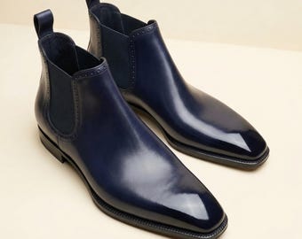 May include: A pair of navy blue Chelsea boots with a high-gloss finish. The boots feature a rounded toe, elastic side panels, and a pull tab at the back. The leather boots are displayed on a neutral background.