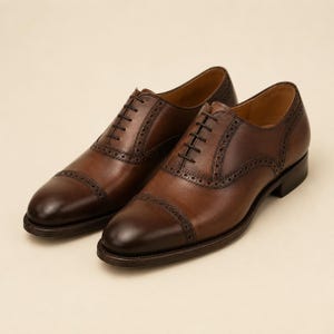 May include: A pair of brown leather oxford shoes featuring a cap toe design with decorative brogue detailing along the seams and eyelet panels. The shoes have a burnished finish at the toe and heel, dark brown laces, and a low stacked heel.