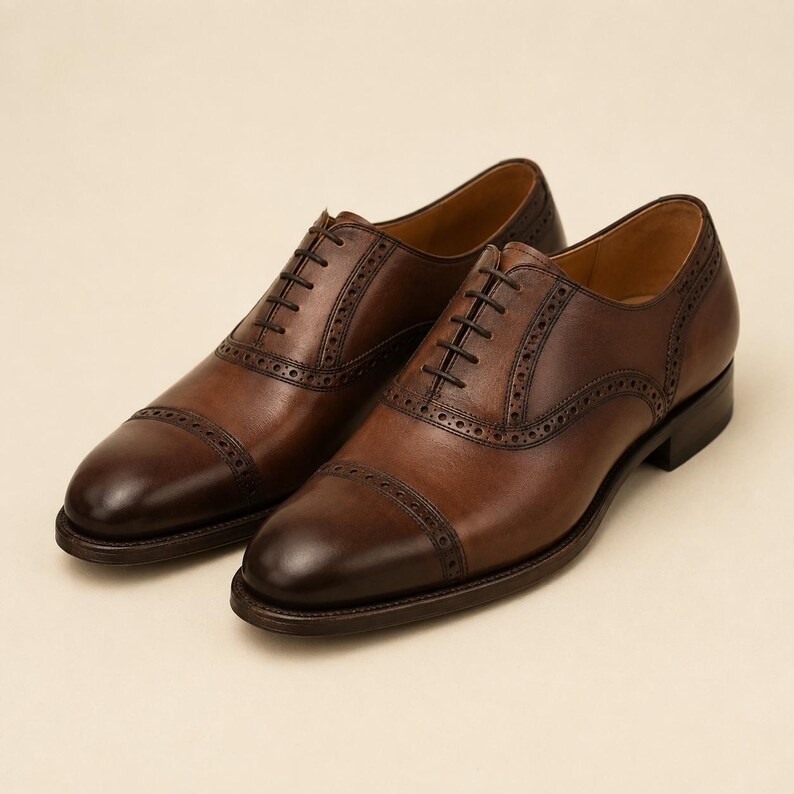 May include: A pair of brown leather oxford shoes featuring a cap toe design with decorative brogue detailing along the seams and eyelet panels. The shoes have a burnished finish at the toe and heel, dark brown laces, and a low stacked heel.