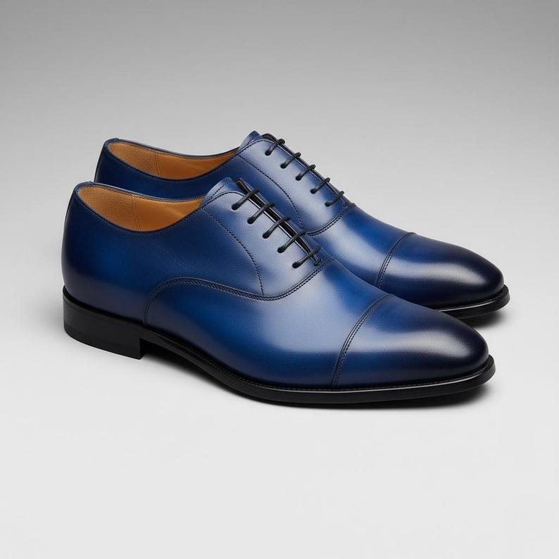 May include: A pair of blue leather dress shoes with black laces and soles. The shoes have a classic Oxford design with a cap toe and a polished finish. The shoes are displayed on a neutral background.