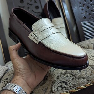 May include: A pair of two-tone penny loafers featuring a dark brown leather body and a contrasting cream-colored leather vamp. The shoes have a classic slip-on design with a horizontal strap across the top and a smooth, dark brown leather sole.