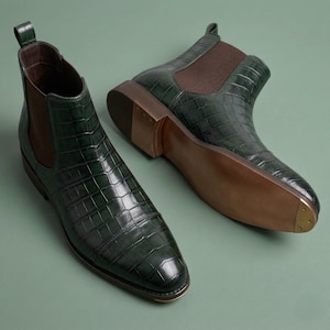 May include: Two dark green Chelsea boots with a crocodile skin texture on a green background. The boots have brown elastic side panels and soles. The heels have a small metal tap.