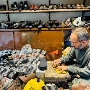 May include: A workshop filled with numerous pairs of black and brown leather shoes arranged on wooden shelves and in plastic packaging. A person is seated at a workbench, using a small tool to work on a piece of yellow leather material.