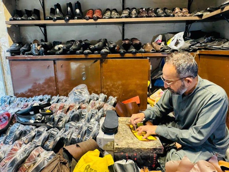 May include: A workshop filled with numerous pairs of black and brown leather shoes arranged on wooden shelves and in plastic packaging. A person is seated at a workbench, using a small tool to work on a piece of yellow leather material.